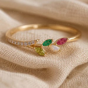 May include: A gold ring featuring a thin band with a row of small clear stones on one side. The focal point consists of four marquise-cut gemstones arranged in a leaf-like cluster, including a green, a pink, a light green, and an iridescent stone.