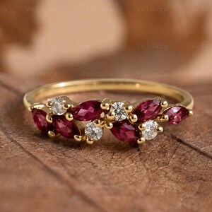 May include: A gold ring featuring a leaf-like design with marquise-cut red gemstones and round, clear gemstones. The ring is set on a brown surface.
