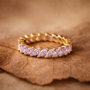 May include: A gold eternity band featuring a continuous row of marquise-cut light pink gemstones set in a metal frame. The ring is circular and displays a repeating pattern of faceted stones that catch the light, creating a uniform appearance.