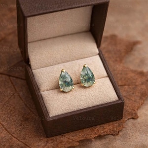 Puede incluir: Un par de pendientes en forma de lágrima con engaste dorado y una piedra moteada verde y blanca. Los pendientes se muestran en una caja de joyería marrón abierta con un interior beige. La palabra "Veloura" está impresa en la caja.