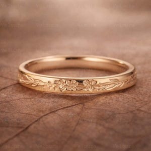 May include: A gold ring with a floral and leaf design. The ring has detailed carvings of flowers and foliage around the band. The ring is presented on a brown surface, highlighting the intricate design and craftsmanship.
