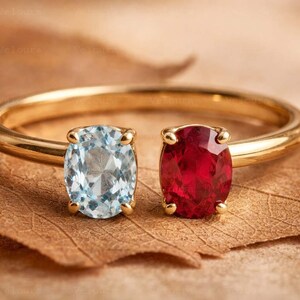 Puede incluir: Un anillo de oro con dos piedras preciosas ovaladas: una aguamarina azul claro y un rubí rojo intenso. El anillo se encuentra sobre una hoja marrón, mostrando el contraste entre el oro y las gemas de colores. El diseño del anillo es abierto.