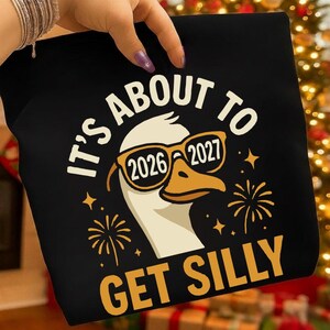 Pode incluir: Camiseta preta com o texto "IT'S ABOUT TO GET SILLY". O design apresenta um ganso usando óculos de sol com os números 2026 e 2027 nas lentes. Fogos de artifício e estrelas também estão incluídos no design.