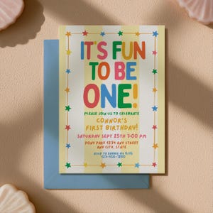Può includere: Un biglietto d'invito di compleanno rettangolare con uno sfondo a righe verticali gialle e bianche, caratterizzato da un bordo di stelle colorate. Il testo recita: IT'S FUN TO BE ONE! PLEASE JOIN US TO CELEBRATE CONNOR'S FIRST BIRTHDAY! SATURDAY SEPT 25TH 3:00 PM PONY PARK 1234 ANY STREET ANY CITY, STATE RSVP TO DEBBIE BY 9/15 123-456-7890. Il biglietto è posizionato su una busta blu.