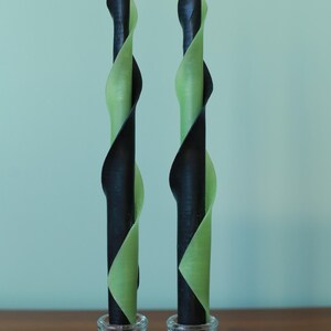 May include: Two tall, slender taper candles featuring a spiraling, dual-color design of matte black and light sage green wax. Each candle is set into a clear, pressed glass candle holder with a tiered, circular base and a white wick.