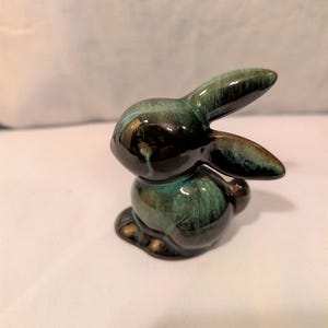 May include: A ceramic rabbit figurine featuring a glossy glaze with a blend of deep black and vibrant teal colours. The rabbit is depicted in a seated position with its ears pointed upward and backward, showcasing a smooth, reflective surface finish.