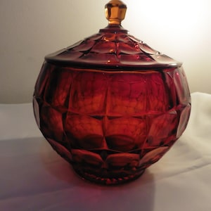 Könnte beinhalten: Eine tiefrote Glasschale mit einem facettierten, geometrischen Rautenmuster auf dem Körper und dem Deckel. Der Behälter hat eine abgerundete Form und ist mit einem kleinen, bernsteinfarbenen, klaren Glasgriff auf dem Deckel versehen.