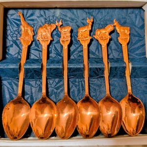 Puede incluir: Un juego de seis cucharas de color cobre con figuras de animales en la parte superior de los mangos. Los animales incluyen una jirafa, un elefante, un ciervo y otros. Las cucharas se exhiben en una caja con un interior azul estampado.