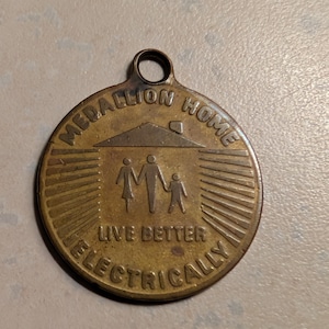 Op de afbeelding: Een ronde, bronskleurige medaillon met de tekst "MEDALLION HOME" en "LIVE BETTER ELECTRICALLY". In het midden een silhouet van een huis en een familie-icoon. Er zit een klein gaatje aan de bovenkant om op te hangen. Diameter ca. 5 cm.