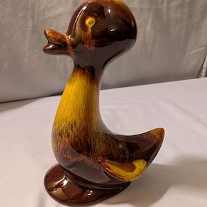 May include: A ceramic duck figurine with a glossy finish. The duck features a brown head and body, transitioning to yellow and brown on the neck and wings. The figurine has an open beak and a rounded base.
