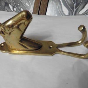 May include: A polished brass wall hook featuring a sculpted horse head at one end and two curved hooks extending from a flat rectangular base. The metal surface is reflective and gold-toned, with two visible screw holes for mounting purposes.