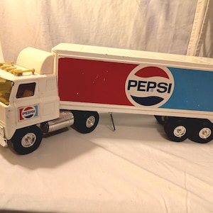 May include: A white toy semi-truck with a trailer featuring the Pepsi logo in red, white, and blue. The truck has black wheels, a silver fuel tank, and a yellow-tinted windshield. The Pepsi logo is also on the truck's door.