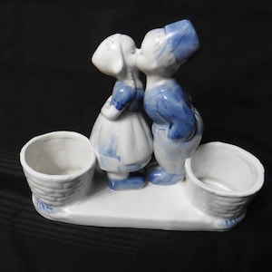 May include: A white ceramic figurine featuring two figures kissing, dressed in blue and white outfits, positioned between two small, round, basket-weave textured containers on a shared base, finished with blue painted accents throughout the piece.