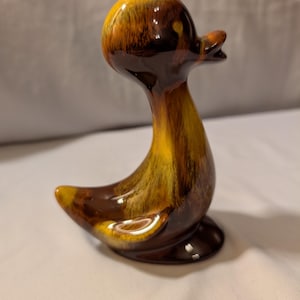 以下が含まれることがあります： 光沢のある仕上がりのセラミック製アヒルの置物。アヒルは茶色、黄色、オレンジ色の混合色です。この置物は、湾曲した体、小さなくちばし、丸いベースが特徴です。装飾品です。