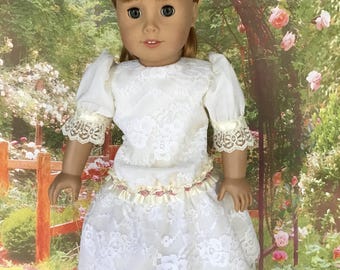 May include: A doll wearing a white lace dress with short sleeves and a ruffled skirt. The dress has a cream-colored ribbon around the waist and lace trim on the sleeves. The doll has blonde hair and is wearing black shoes. The doll is standing on a white base.