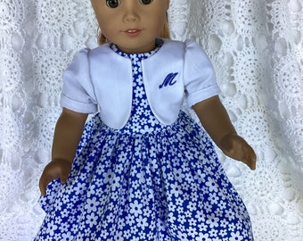 May include: A doll wearing a white cardigan with a blue "M" embroidered on the front, over a blue dress with a white floral pattern. The doll has brown hair and brown eyes.