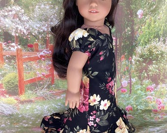 May include: A doll with long, dark hair and a red flower in her hair is wearing a black floral print dress. The dress features a ruffled hem and is adorned with pink, yellow, and white flowers. The doll is posed in front of a garden backdrop.