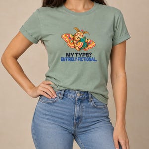 May include: A sage green t-shirt featuring a graphic of a moth holding a book with a small heart above its head. Below the moth, the text reads "MY TYPE? ENTIRELY FICTIONAL." in black and blue lettering. The shirt has short sleeves.
