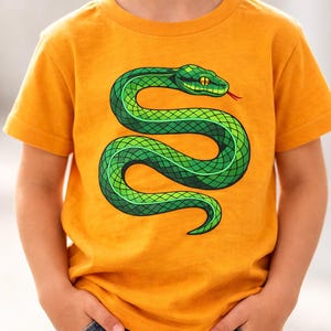 Peut inclure: Un t-shirt à manches courtes jaune moutarde arborant un grand motif détaillé représentant un serpent vert enroulé en forme de S. Le serpent présente un motif d'écailles texturé, un œil jaune vif et une petite langue rouge bifide.