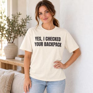 May include: A cream-colored short-sleeve t-shirt featuring the black text YES, I CHECKED YOUR BACKPACK printed in a bold, sans-serif font across the chest. The garment has a crew neckline and a relaxed fit, paired with light blue denim jeans.