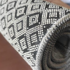 May include: Rolled-up black and white patterned rug. The rug features a repeating geometric diamond design. The edges of the rug are a solid gray color. The rug is made of a woven material.