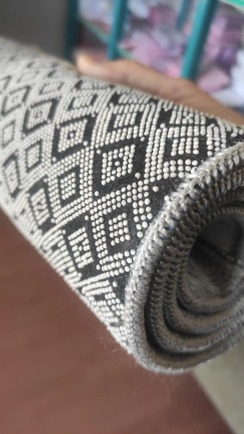 May include: Rolled-up black and white patterned rug. The rug features a repeating geometric diamond design. The edges of the rug are a solid gray color. The rug is made of a woven material.