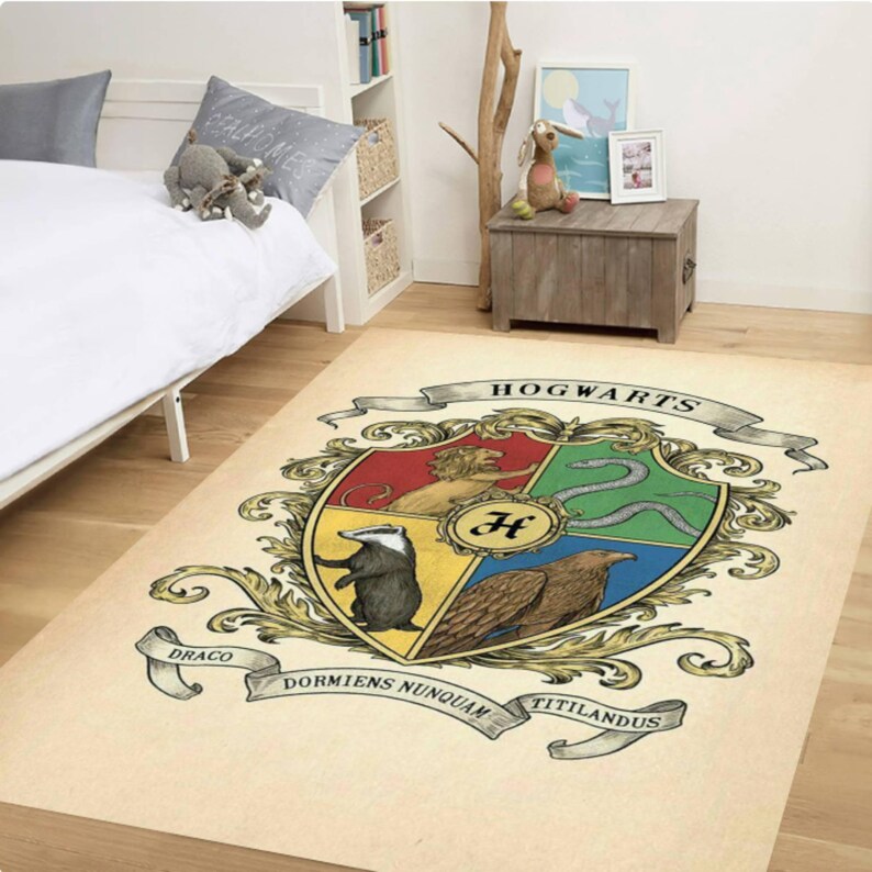 May include: A rectangular rug featuring the Hogwarts crest, with a cream-colored background. The crest includes a lion, snake, badger, and eagle, with the word "Hogwarts" across the top. The rug is placed in a bedroom setting.