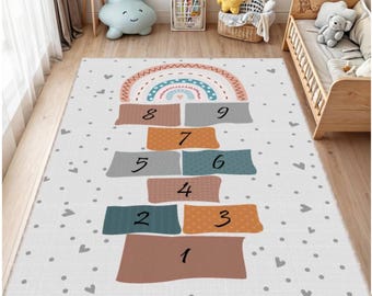 May include: A rectangular children's play rug featuring a hopscotch design. The rug has a white background with gray hearts and polka dots. The hopscotch squares are in shades of brown, orange, gray, and teal, with a rainbow at the top.