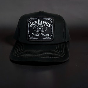 May include: A black baseball cap featuring a rectangular patch on the front. The patch displays the white text Jack Daniel's Old No. 7 Brand Field Tester with decorative scrollwork borders. The cap has a structured crown and a flat brim with stitching.
