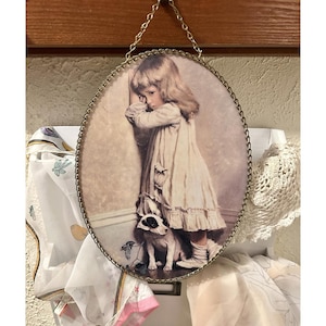 May include: Oval-shaped framed print featuring a young child in a white dress, leaning against a wall, with a small dog at their feet. The frame has a decorative gold-colored edge and is suspended by a gold chain.
