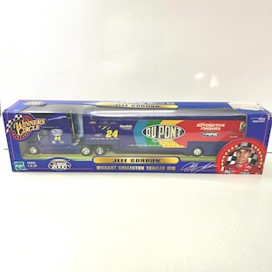 May include: A diecast collector trailer rig featuring Jeff Gordon. The blue truck and trailer have the number 24, along with sponsor logos including DuPont and Pepsi. The packaging includes the Winners Circle logo and a picture of Jeff Gordon.