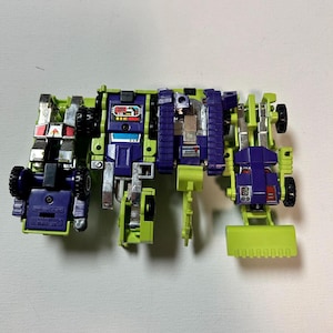 May include: A vintage toy robot in a construction vehicle configuration. The toy is primarily green, purple, and silver, with black wheels and a front scoop. The robot is made of multiple articulated parts.