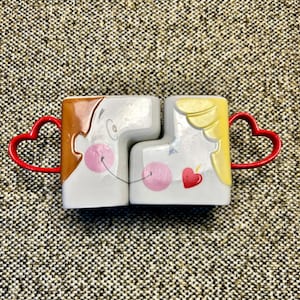 May include: Two ceramic mugs designed to fit together, depicting a couple kissing. One mug has brown hair, the other has yellow hair. Both mugs have red heart-shaped handles and pink blush marks.