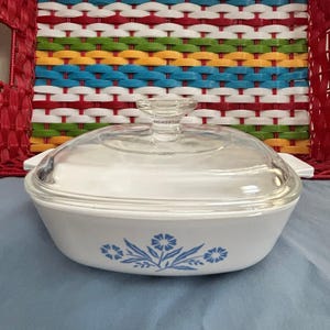 May include: White oval casserole dish with a clear glass lid. The dish features a blue floral design on the front. The handles are white. The background includes a colorful woven basket.