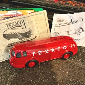 May include: A red Texaco 1934 Diamond T Tanker toy bank with black wheels and white lettering. The bank has a slot for coins and is accompanied by vintage printed materials. The word "Texaco" is prominently displayed.