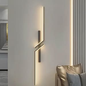 May include: Modern wall sconce with a linear design. The sconce features a vertical light strip and a geometric intersection of lines, emitting a warm glow. The fixture is mounted on a neutral-colored wall, creating a minimalist aesthetic.