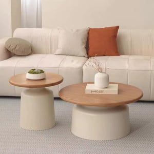 May include: Two modern coffee tables with a light wood top and a cream-colored base. One table is smaller and round, while the other is larger. A white sofa and decorative pillows are in the background.