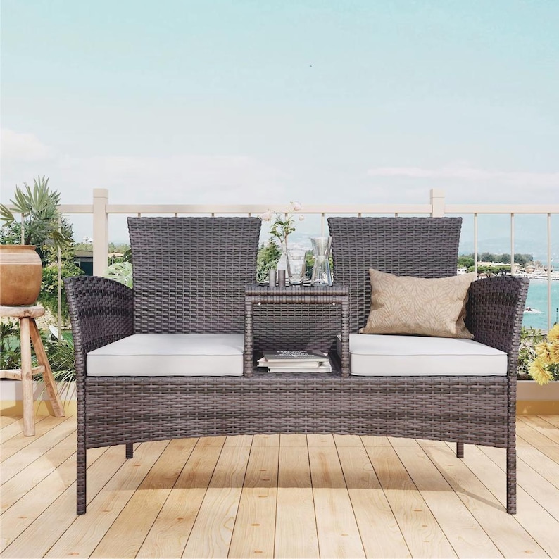 Puede incluir: Un sof&aacute; de dos plazas de mimbre gris para exteriores con una mesa auxiliar incorporada. El sof&aacute; tiene cojines blancos y un coj&iacute;n decorativo. El escenario es en una terraza de madera con vistas al mar.