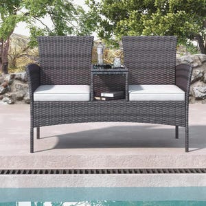 Puede incluir: Un sof&aacute; de exterior de mimbre gris oscuro con una mesa auxiliar incorporada. El sof&aacute; tiene dos asientos con cojines blancos y un ribete negro. Una peque&ntilde;a mesa de cristal est&aacute; entre los asientos, con una botella y un peque&ntilde;o objeto decorativo. Dos libros est&aacute;n en el estante.