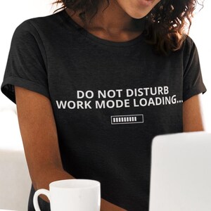 Pode incluir: Uma mulher usando uma camiseta cinza escura com o texto "DO NOT DISTURB WORK MODE LOADING..." em branco. Ela está sorrindo e usando um laptop branco. Uma caneca branca está na mesa na frente dela.