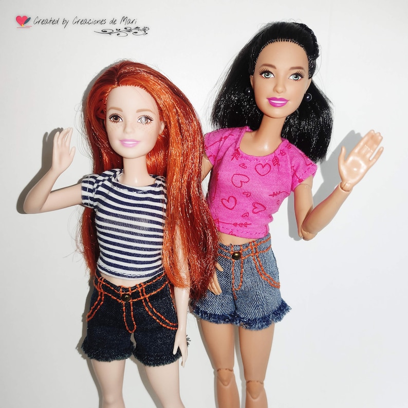 May include: Two dolls are featured, one with long red hair and a striped shirt, the other with black hair and a pink heart-printed top. Both dolls wear denim shorts with orange stitching. The dolls are posed with their hands raised.