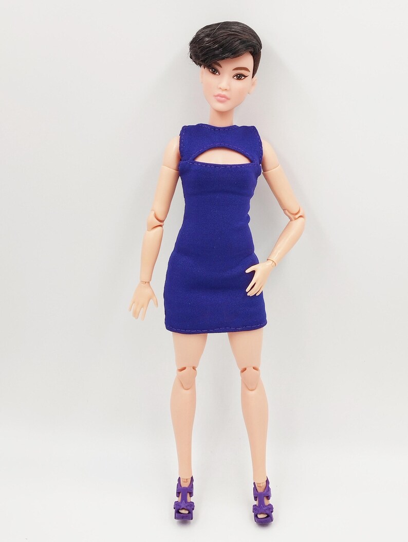 May include: A fashion doll in a royal blue mini-dress with a keyhole detail. The doll has short, dark hair and is wearing purple high-heeled shoes. The doll is set against a plain white backdrop.