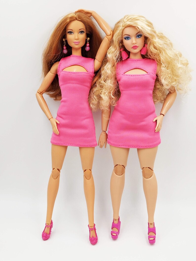 May include: Two fashion dolls, each in a bright pink dress with a keyhole neckline. One doll has auburn hair, the other blonde. Both wear matching pink earrings and shoes. The dolls stand on a white surface. The dresses are a vibrant, eye-catching colour.