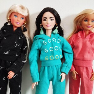 K&ouml;nnte beinhalten: Drei Puppen in passenden Outfits. Eine Puppe tr&auml;gt einen schwarzen Trainingsanzug mit wei&szlig;en astrologischen Symbolen. Eine andere Puppe tr&auml;gt einen t&uuml;rkisfarbenen Trainingsanzug mit dem Wort "GOOD" wiederholt. Die dritte Puppe tr&auml;gt einen rosa Trainingsanzug.