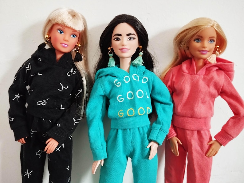 K&ouml;nnte beinhalten: Drei Puppen in passenden Outfits. Eine Puppe tr&auml;gt einen schwarzen Trainingsanzug mit wei&szlig;en astrologischen Symbolen. Eine andere Puppe tr&auml;gt einen t&uuml;rkisfarbenen Trainingsanzug mit dem Wort "GOOD" wiederholt. Die dritte Puppe tr&auml;gt einen rosa Trainingsanzug.