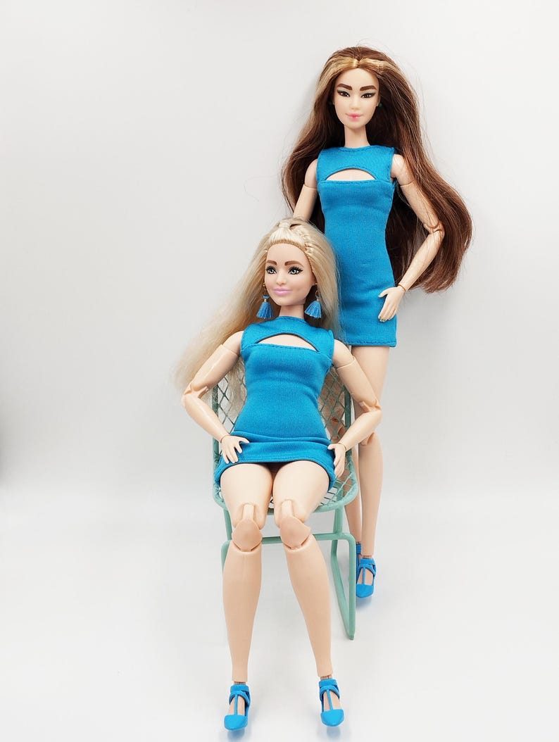 May include: Two fashion dolls in matching blue dresses with keyhole necklines. One doll sits on a pale green chair, the other stands behind. Both wear blue high-heeled shoes. The dolls have long hair, one blonde and one brunette. The dolls are a fashion item.