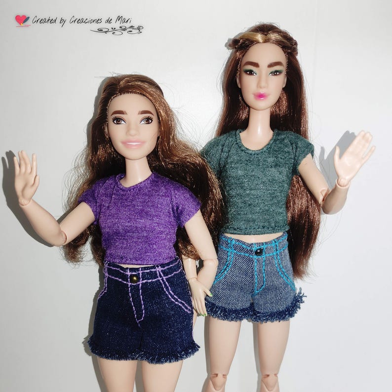 May include: Two dolls wearing matching outfits. One doll is wearing a purple crop top and denim shorts with white stitching. The other doll is wearing a green crop top and denim shorts with blue stitching. Both dolls have long brown hair.
