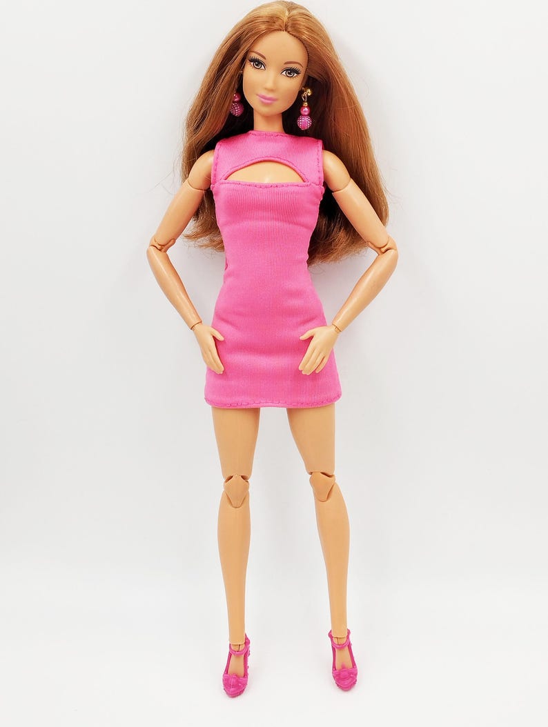 May include: A fashion doll with long auburn hair, wearing a bright pink dress with a keyhole neckline. The doll is accessorized with pink earrings and matching pink heels. The doll is standing with arms at her sides.