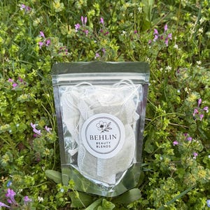 Könnte beinhalten: Ein klarer Plastikbeutel gefüllt mit weißen Teebeuteln, mit der Aufschrift "BEHLIN BEAUTY BLENDS" und einem floralen Logo. Der Beutel liegt vor einem Hintergrund aus grünem Gras und kleinen lila und weißen Wildblumen.