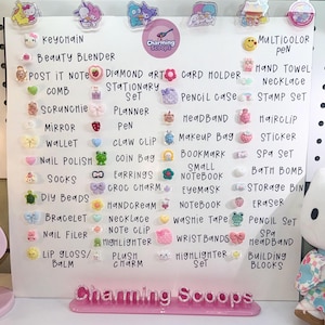 Op de afbeelding: Een wit displaybord met diverse kleine, kleurrijke accessoires, waaronder sleutelhangers, scrunchies en stickers. Het bord heeft handgeschreven labels en de tekst "Charming Scoops."